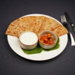 Tasty Indian paratha served with pickle and yogurt on a white plate with cutlery.